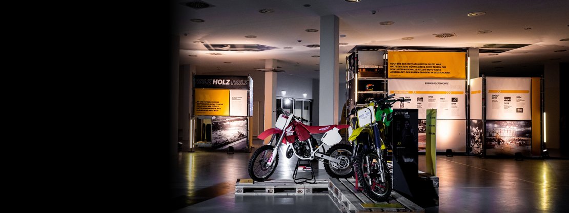VISUELL Szenografie: Ausstellung: Zwei historische Motorräder sind auf Paletten erhöht präsentiert; dahinter die modularen Gerüstsysteme mit Bannern, Texten und leuchtenden Elementen.