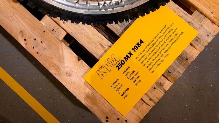 VISUELL Szenografie: Ausstellung: Detailansicht einer gelben Objektbeschirftung mit technischen Daten zur KTM 250 MX von 1984; daneben das Rad eines Motorrads auf Holzpaletten.