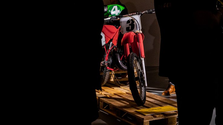 VISUELL Szenografie: Ausstellung: Ein rotes Motorrad mit Startnummer „3“ ist auf Paletten erhöht präsentiert; Besucherinnen und Besucher betrachten das Exponat im Halbdunkel.