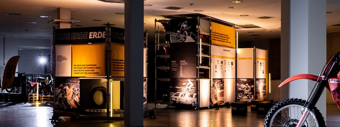 VISUELL Szenografie: Ausstellung: Offene Raumansicht mit Gerüstkuben, Bannern und Fotos; ein rotes Motorrad im Vordergrund bildet den Auftakt zur Inszenierung.