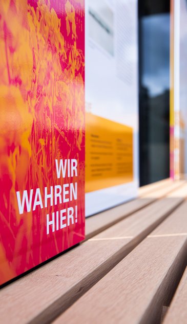 VISUELL Szenografie: Wahre Wunderbänke: Nahaufnahme einer Sitzbank mit Informationstafel mit plakatives Bildmotiv in magenta und orange mit der Aufschrift WIR WAHREN HIER!