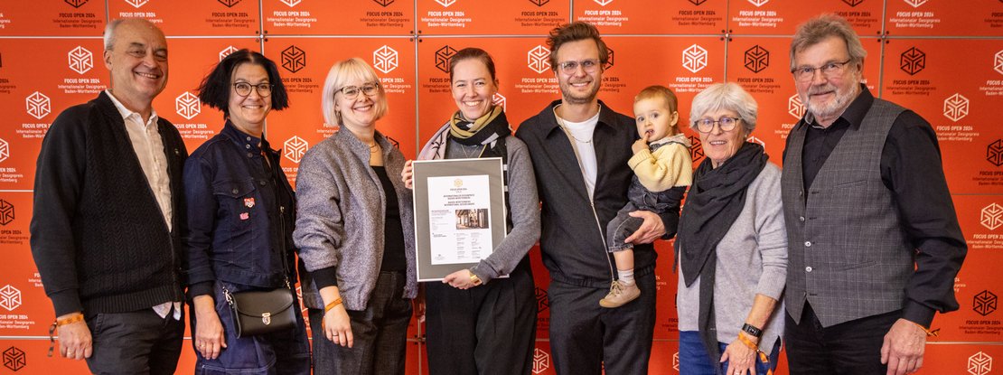 VISUELL Blogbeitrag: FOCUS OPEN Award 2024 Gold: Sieben Personen aus dem Projektteam posieren mit der Urkunde des FOCUS OPEN Award 2024 vor einem Fotohintergrund mit dem Logo des Internationalen Designpreis Baden-Württemberg