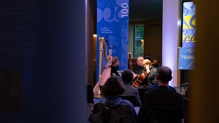 VISUELL Szenografie: Jubiläumsausstellung: Einblick in die Eröffnungsveranstaltung über die Schultern des Publikums auf die Musikerinnen und Musiker des Orchesters