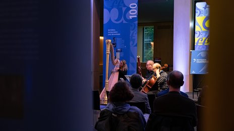 VISUELL Szenografie: Jubiläumsausstellung: Einblick in die Eröffnungsveranstaltung über die Schultern des Publikums auf die Musikerinnen und Musiker des Orchesters