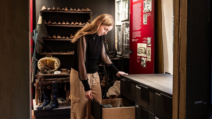 VISUELL Scenography: Permanent exhibition at Hornmoldhaus: Visitor opens a drawer of a black display cabinet with integrated exhibits. In the background, a historical workshop setting and a red information board on the town's history.