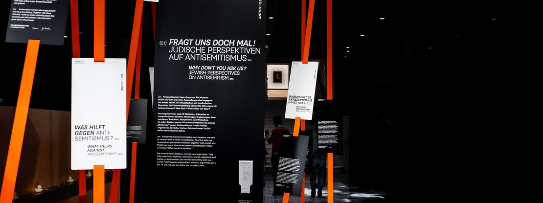VISUELL Szenografie: Sonderausstellung: Anti Anti 2.0: Vertikale Installation mit leuchtend orangefarbenen Bändern; daran befestigte, schwarze und weiße Informationstafeln enthalten Typografie