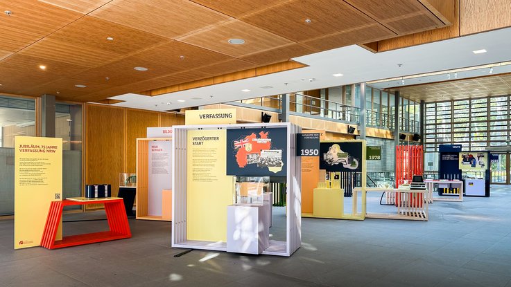 VISUELL Szenografie: Wanderausstellung „Jubiläum. 75 Jahre Verfassung NRW“ für die Stiftung Haus der Geschichte Nordrhein-Westfalen VISUELL Szenografie: Wanderausstellung: Gesamtansicht der Ausstellung in einem modernen Foyer. Mehrere Themenstationen in klarer Farbstruktur, im Vordergrund der Bereich „Verfassung – Verzögerter Start“.