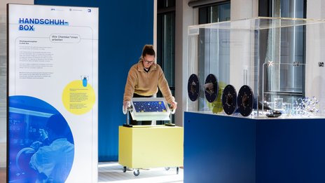 VISUELL Szenografie: Wissenschaftsausstellung: Eine Besucherin bedient eins der zwei interaktiven Exponate in der Ausstellung „Was kann Katalyse?“, daneben befindet sich eine Informationstafel mit Text und Bild