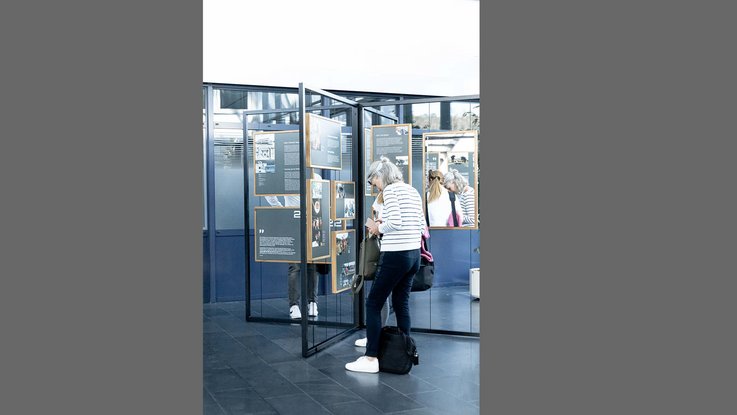 VISUELL Szenografie: Jubiläumsausstellung: Verschiedene Personen beschäftigen sich mit den Inhalten der Ausstellung; eine Tafel ist mit Spiegelfolie ausgestattet, sodass man zwei Personen ebenfalls in dieser Spiegelung sieht.