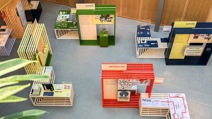 VISUELL Szenografie: Wanderausstellung „Jubiläum. 75 Jahre Verfassung NRW“ für die Stiftung Haus der Geschichte Nordrhein-Westfalen VISUELL Szenografie: Wanderausstellung: Vogelperspektive auf den Ausstellungsaufbau mit Modulen in Grün, Gelb, Rot und Blau. Themenbereiche wie Umwelt, Mitsprache, Europa und Datenschutz sind deutlich erkennbar.