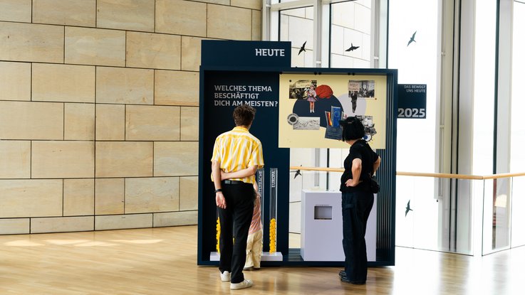 VISUELL Szenografie: Wanderausstellung „Jubiläum. 75 Jahre Verfassung NRW“ für die Stiftung Haus der Geschichte Nordrhein-Westfalen VISUELL Szenografie: Wanderausstellung: Zwei Personen betrachten das Ausstellungselement „Heute – Welches Thema beschäftigt dich am meisten?“. Gelb-blaue Gestaltung mit Themenröhren zur Publikumsbeteiligung.