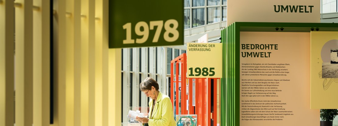 VISUELL Szenografie: Wanderausstellung: Nahaufnahme der Themenmodule Umwelt und Verfassungsänderung. Im Vordergrund gelbe Lamellenstruktur mit den Jahreszahlen 1978 und 1985, eine Besucherin liest in einem Buch.