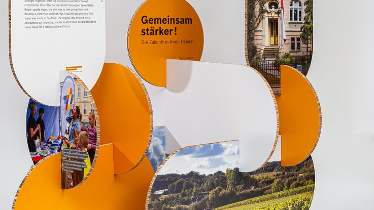 VISUELL Szenografie: Wanderausstellung Robert Bosch Stiftung GmbH: Detailansicht einer organisch geformten Ausstellungsstation aus orangefarbenen und weißen Wabenplatten; integrierte Bildtafeln zeigen Begegnungsszenen und Landschaften, darüber der Schriftzug „Gemeinsam stärker! Die Zukunft in Ihren Händen.“