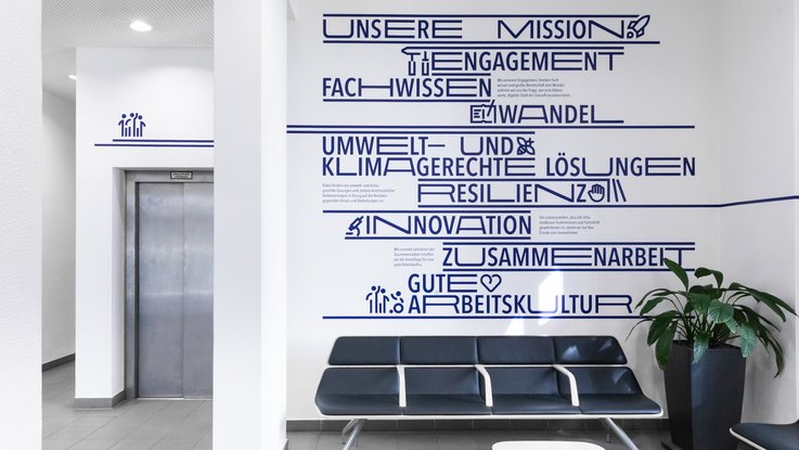 VISUELL Kommunikationsdesign: Wandgrafiken Landeshauptstadt Stuttgart: Wartebereich mit Sitzmöbeln und zentral platzierter Wandgrafik zur Mission und den Werten der Institution.