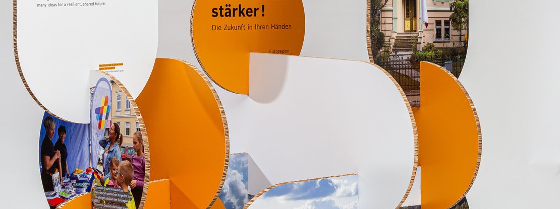 VISUELL Scenography: Robert Bosch Stiftung GmbH travelling exhibition: a close-up of an organically shaped exhibition stand made of orange and white honeycomb panels; integrated display panels show scenes from events and landscapes, with lettering above that ends with “…stronger! The future is in your hands”.