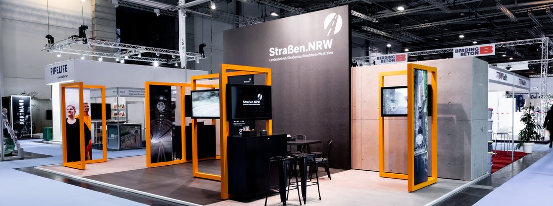 VISUELL Scenography: Straßen.NRW exhibition stand at InfraTech 2026: Photographed from the corner of the stand: a striking orange frame structure divides the 50-square-metre stand area into various themed zones featuring integrated screens and information panels.