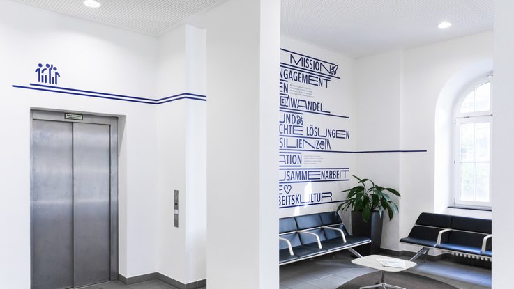 VISUELL Kommunikationsdesign: Wandgrafiken Landeshauptstadt Stuttgart: Wartebereich mit Aufzug, Sitzmöbeln und umlaufender Liniengrafik, die Räume visuell verbindet.
