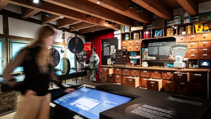 VISUELL Scenography: Permanent exhibition Hornmoldhaus: Exhibition room with historic merchant's cabinet and numerous drawers, supplemented by a shop scale and goods packaging. In the foreground, an interactive media table with touch display; in the background, round portrait panels and thematic information boards in the half-timbered room.