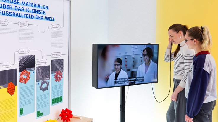 VISUELL Szenografie: Wissenschaftsausstellung: Zwei Besucherinnen stehen vor einer Bildschirmstele und verfolgen den abgespielten Film, eine von ihnen hält einen Handkopfhörer an ihr Ohr. Daneben befindet sich ein Plakat mit Informationen und mehrere Modelle von Molekülstrukturen von unterschiedlichen Materialien.