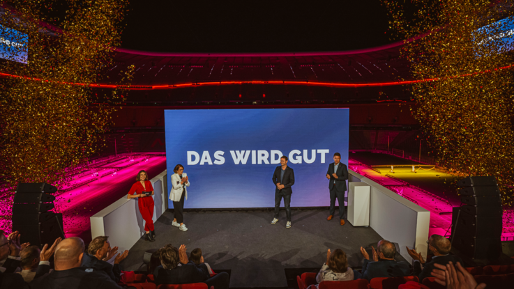 VISUELL Szenografie: Event „NEXT LEVEL“: Vier Personen stehen vor der großen LED-Leinwand auf der „Das wird gut“ zu lesen ist, zum feierlichen Abschluss des Events wird links und rechts von der Bühne goldenes Konfetti in die Luft geschossen.