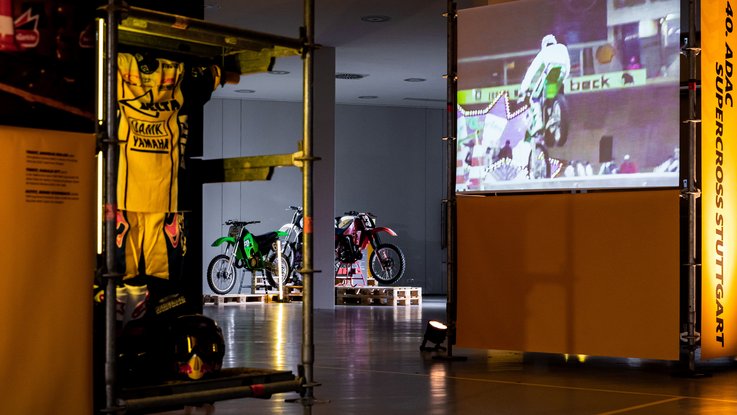 VISUELL Szenografie: Ausstellung: Blick durch ein Gerüstmodul mit aufgehängtem gelbem Rennanzug; im Hintergrund auf Paletten inszenierte historische Motorräder, rechts eine große Projektion mit Rennszenen.