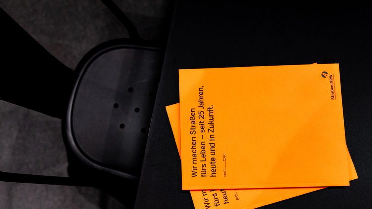 VISUELL Scenography: Straßen.NRW stand at InfraTech 2026: A close-up of the anniversary brochure designed by VISUELL, “We build roads for life – for 25 years”, in bright orange, resting on a simple black display unit.