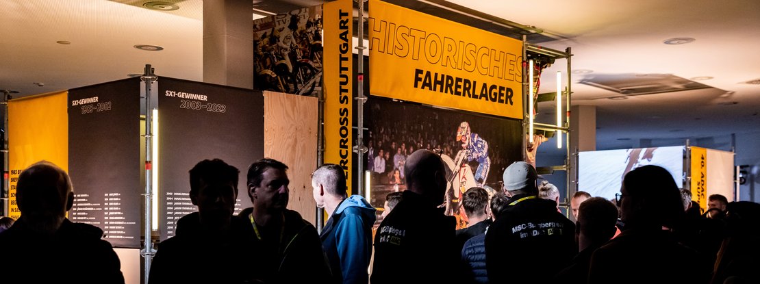 VISUELL Szenografie: Ausstellung: Besucherinnen und Besucher versammeln sich vor den großen Tafeln mit Zeitstrahl und Siegerlisten; darüber prangt ein gelbes Banner mit der Aufschrift „Historisches Fahrerlager“.