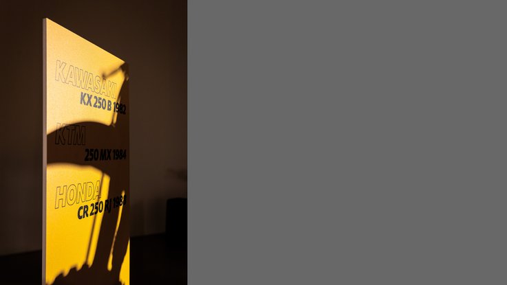 VISUELL Szenografie: Ausstellung: Nahaufnahme einer gelben Infotafel mit aufgedruckten Modellnamen; Licht und Schattenwurf eines Motorrads schaffen eine spannende Atmosphäre.