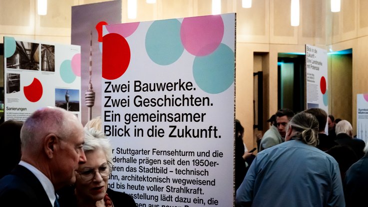 VISUELL Szenografie: Jubiläumsausstellung: 70 Jahre Fernsehturm und Liederhalle: Besucher:innen stehen vor einer großformatigen Ausstellungsgrafik mit historischen Fotografien und Texten zur Entstehung des Fernsehturms.