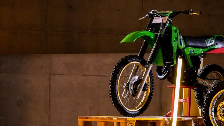 VISUELL Szenografie: Ausstellung: Ein grünes Motorrad wird auf Paletten und mit Lichtstelen in Szene gesetzt; Betonwand und Bodenmarkierungen verstärken das industrielle Ambiente.