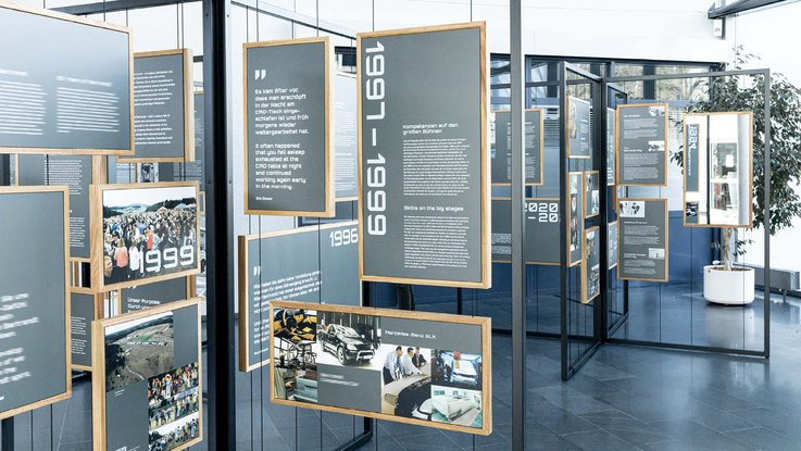 VISUELL Szenografie: Jubiläumsausstellung: Aufnahme der Rahmenkonstruktion aus Metall mit Stahlseilen und vielerlei Holzrahmen mit unterschiedlich gestalteten Informationstafeln mit Jahreszahlen, Bildern, Zitaten und Texten.