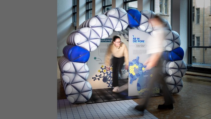 VISUELL Szenografie: Eine Besucherin steht in einem begehbaren, aufgeblasenen Modell einer Pore in der Wissenschaftsausstellung „Was kann Katalyse?“. Die Struktur des Modells besteht aus Molekülen und ist als Bogen aufgebaut. Im Inneren der Pore befindet sich mehrere Informationstafeln. Eine weitere Besucherin läuft im Vordergrund an dem Exponat vorbei.