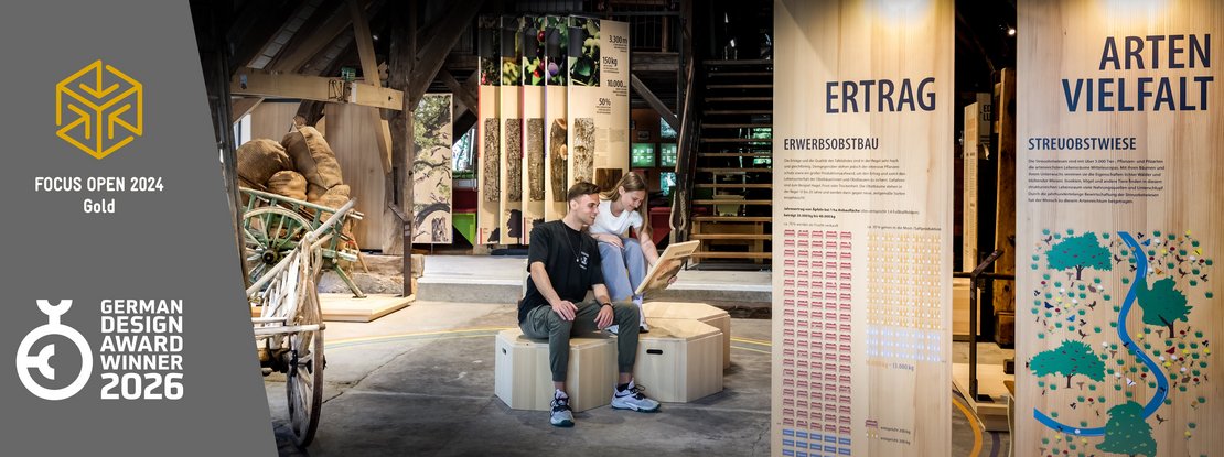 VISUELL Szenografie: Obstbaumuseum: Zwei Besucher sitzen in der Mitte der Ausstellung und betrachten eine Informationstafel in ihren Händen. Links neben dem Bild stehen Logos von Auszeichnungen, die das Projekt erhalten hat: Focus Open 2024 in Gold und German Design Award Winner 2026