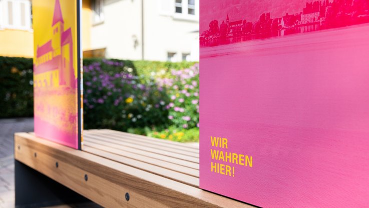 VISUELL Szenografie: Wahre Wunderbänke: Rückseite einer Sitzbank mit Informationstafeln in magenta und gelb mit der Aufschrift WIR WAHREN HIER!