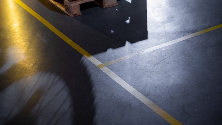 VISUELL Szenografie: Ausstellung: Gelbe Bodenmarkierungen kreuzen sich auf dem grauen Boden; Schatten von Motorrädern und Paletten schaffen eine grafische Wirkung.