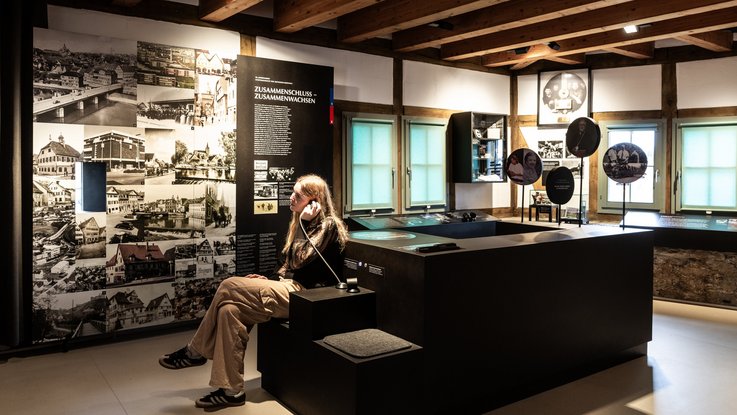 VISUAL Scenography: Permanent exhibition at Hornmoldhaus: Historical exhibition space with exposed wooden beams and a central black exhibition table. A visitor uses an audio station with headphones. Large-format text panel "Merger – Growing Together" and historical photo collage structure the chronological history of the city.