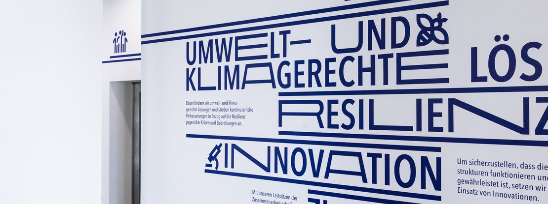 VISUELL Kommunikationsdesign: Wandgrafiken Landeshauptstadt Stuttgart: Großformatige Wandgrafik mit typografisch hervorgehobenen Begriffen wie Umwelt- und klimagerechte Lösungen, Resilienz und Innovation in tiefem Dunkelblau auf weißer Wand.