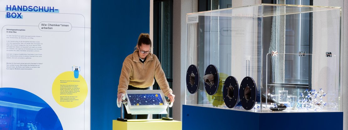 VISUELL Szenografie: Wissenschaftsausstellung: Eine Besucherin bedient eins der zwei interaktiven Exponate in der Ausstellung „Was kann Katalyse?“, daneben befindet sich eine Informationstafel mit Text und Bild
