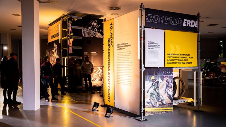VISUELL Szenografie: Ausstellung: Besuchergruppe vor den großformatigen Bannern; helle Projektionsflächen und Texte in Schwarz-Gelb vermitteln historische Hintergründe.