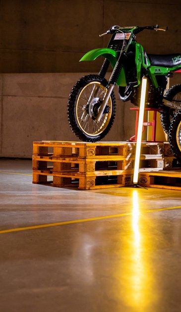 VISUELL Szenografie: Ausstellung: Ein grünes Motorrad wird auf Paletten und mit Lichtstelen in Szene gesetzt; Betonwand und Bodenmarkierungen verstärken das industrielle Ambiente.