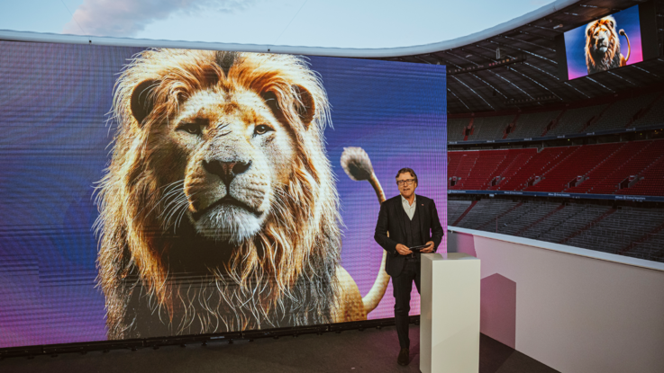 VISUELL Szenografie: Event „NEXT LEVEL“: Eine Person steht hinter einem Rednerpult vor der großen LED-Leinwand auf der Bühne. Im Hintergrund sieht man die leeren ränge der Allianz Arena und und die Hauptstadions-LED, auf beiden LED-Wänden ist ein Löwe zu sehen.