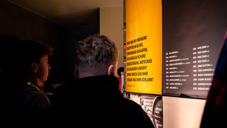 VISUELL Szenografie: Ausstellung: Besuchende lesen aufmerksam die großformatigen Texttafeln mit Zitaten und Siegerlisten; die klare Typografie lenkt den Blick auf die Inhalte.