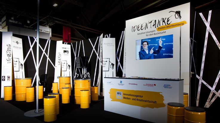 VISUELL Szenografie: Messestand Ideentanke MFG: Gesamtüberblick über den Messestand mit gelben Sitztonne, typografischen Bannern und Screens