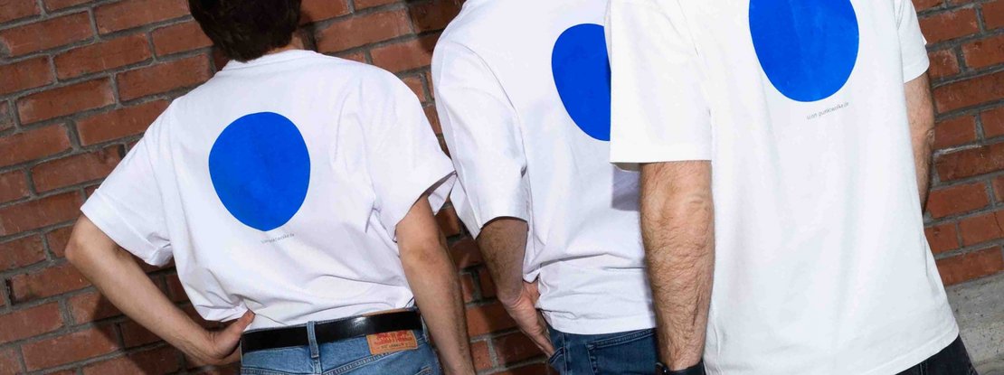 VISUELL Kommunikationsdesign: Corporate Design punktWOLKE: Nahaufnahme von drei Männern, die mit vor einer Backsteinwand stehen und weiße T-Shirts mit einem leuchtend blauen Punkt auf dem Rücken tragen