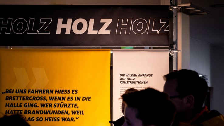 VISUELL Szenografie: Ausstellung: Großaufnahme eines Banners mit der Aufschrift „HOLZ HOLZ HOLZ“; darunter ein Zitatbanner und erläuternde Texte über die Anfänge der Konstruktionen.