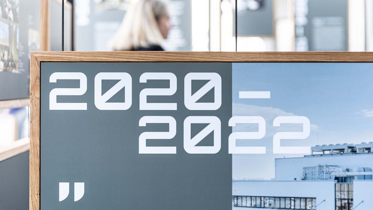 VISUELL Szenografie: Jubiläumsausstellung: Plakative Jahreszahlen „2020–2022“ in der Hausschrift auf einer Informationstafel mit Bildmotiv eines Gebäudes.