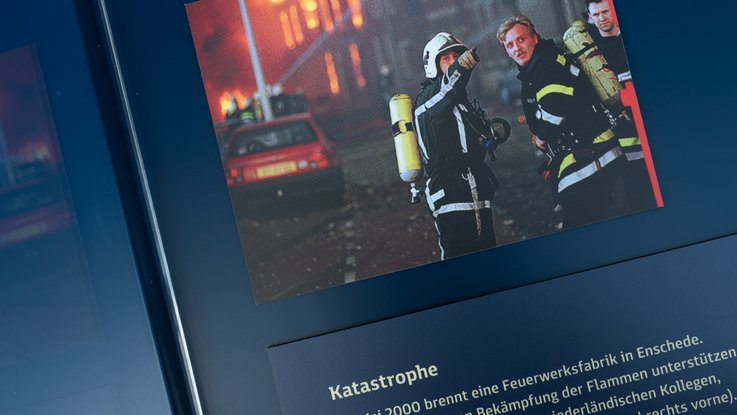 VISUELL Szenografie: Wanderausstellung „Jubiläum. 75 Jahre Verfassung NRW“ für die Stiftung Haus der Geschichte Nordrhein-Westfalen VISUELL Szenografie: Wanderausstellung: Nahaufnahme einer Infotafel mit historischem Foto von Feuerwehrleuten bei einem Großbrand. Überschrift „Katastrophe“, Text zur Explosion in Enschede 2000.