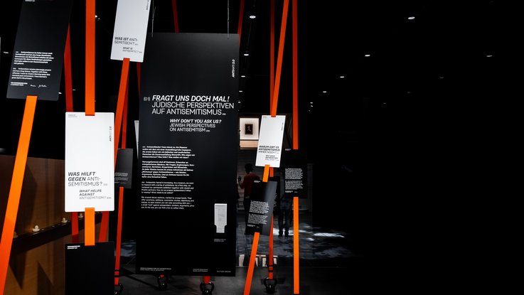 VISUELL Szenografie: Sonderausstellung: Anti Anti 2.0: Vertikale Installation mit leuchtend orangefarbenen Bändern; daran befestigte, schwarze und weiße Informationstafeln enthalten Typografie
