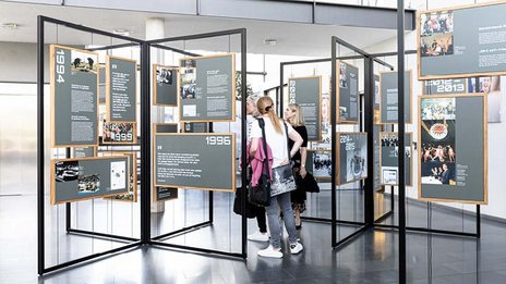 VISUELL Szenografie: Jubiläumsausstellung: Drei Besucherinnen stehen inmitten der Ausstellung und betrachten die Inhalte.