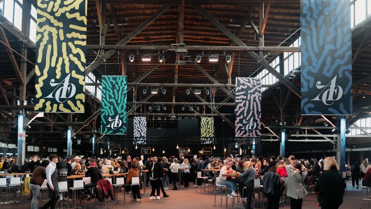 VISUELL Blogbeitrag: ADC Talent Festival 2025 „Goldenes Konfetti – Silberner Nagel“ VISUELL Blogbeitrag: ADC Talent Award 2025: Blick in die Festivalhalle mit bunten Bannern und Besucher:innen, Atmosphäre des Kreativfestivals in Hamburg.